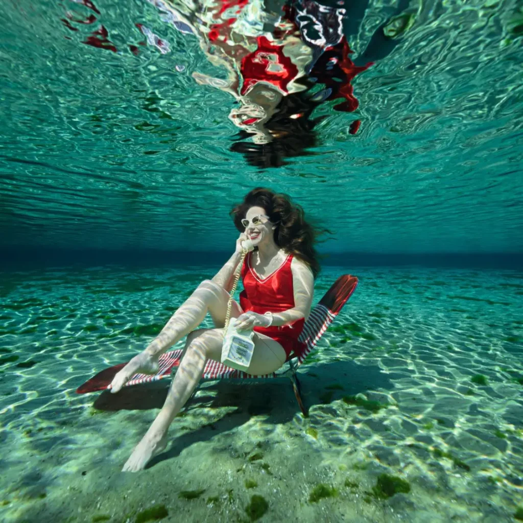 Shannon Scott sitting gracefully underwater taking a phone call in a bright red bathing suit surrounded by soft light and serene ocean blues.