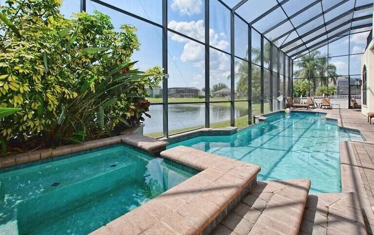 Screened-in pool and spa area with a paver deck, overlooking a lake, surrounded by lush tropical landscaping including palm trees and shrubs, under a bright blue sky with scattered clouds.
