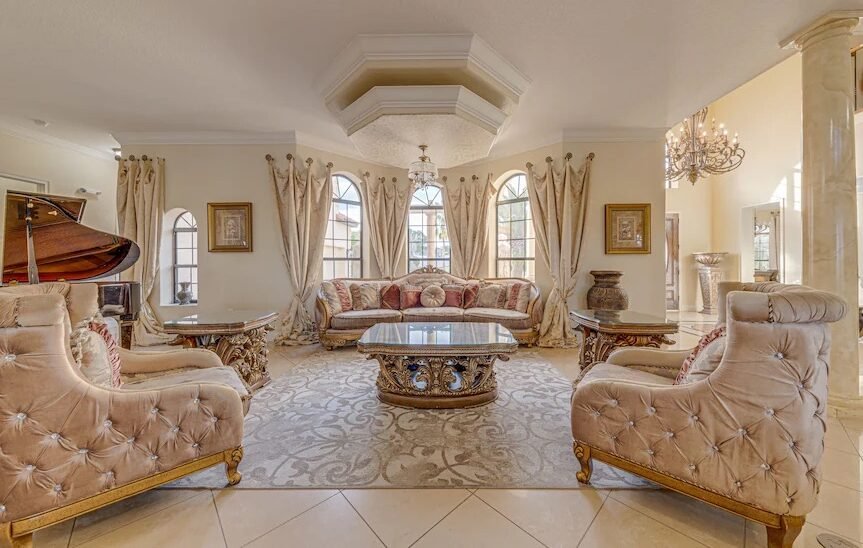 Elegant formal living room with tufted cream sofas and armchairs arranged around a carved wood coffee table, arched windows with draped curtains, a crystal chandelier, ornate crown molding, marble columns, and a grand piano in the corner.